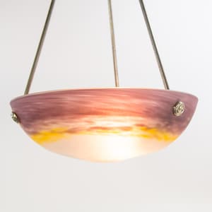 Pu&ograve; includere: Una lampada a sospensione con un paralume a forma di ciotola. Il paralume presenta una miscela di colori, tra cui viola, arancione e giallo, che ricordano un tramonto. La luce &egrave; sospesa da tre aste metalliche e presenta dettagli metallici decorativi.