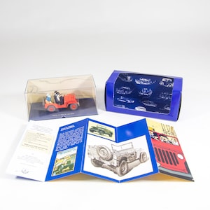 May include: A red toy jeep in a clear plastic display box with a blue box next to it. The jeep has two figures in the front seat. The blue box has a white background with a blue pattern of cars and trucks. The blue box is open and the inside is visible. The toy jeep is on a white background. The toy jeep is part of a set that includes a booklet with images of the jeep and other vehicles. The booklet has the words 'The American Jeep' printed on the cover.