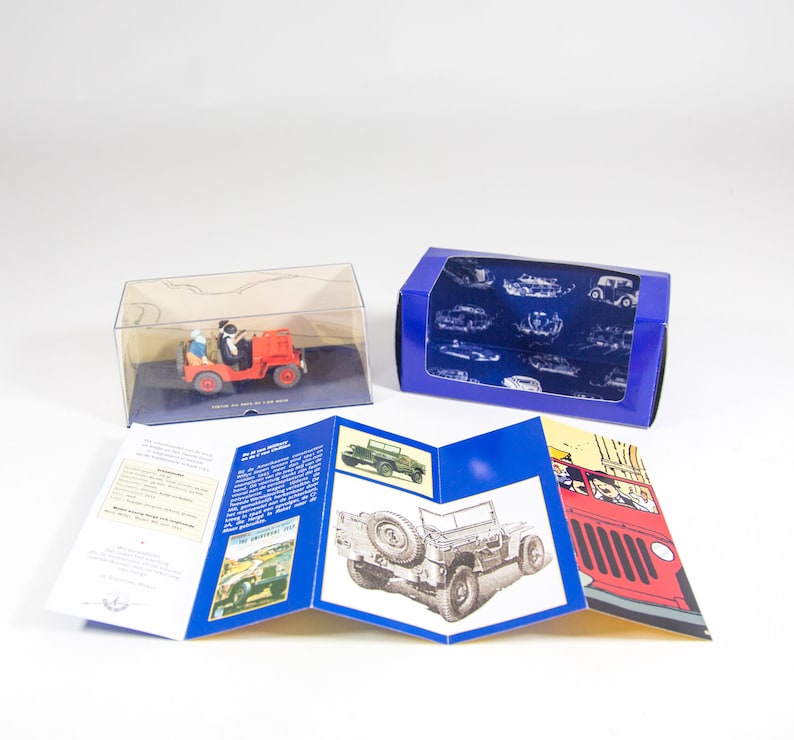 May include: A red toy jeep in a clear plastic display box with a blue box next to it. The jeep has two figures in the front seat. The blue box has a white background with a blue pattern of cars and trucks. The blue box is open and the inside is visible. The toy jeep is on a white background. The toy jeep is part of a set that includes a booklet with images of the jeep and other vehicles. The booklet has the words 'The American Jeep' printed on the cover.