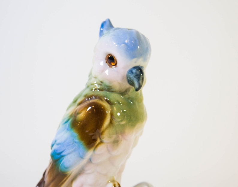 May include: A ceramic figurine of a blue, green, and brown parrot with a white chest and a black beak. The parrot is perched on a branch.