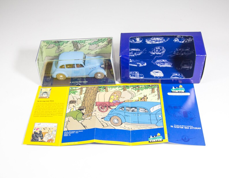 May include: A blue toy car in a clear plastic box with a comic book style illustration of the car on the box. The car is also pictured in a comic book style illustration on a yellow and blue folded card with the text "De Scepter Van Ottokar" on the card.