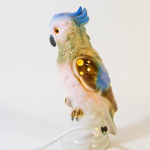 May include: A ceramic night light shaped like a parrot with blue, green, brown, and pink accents. The parrot is perched on a white base with a cord.