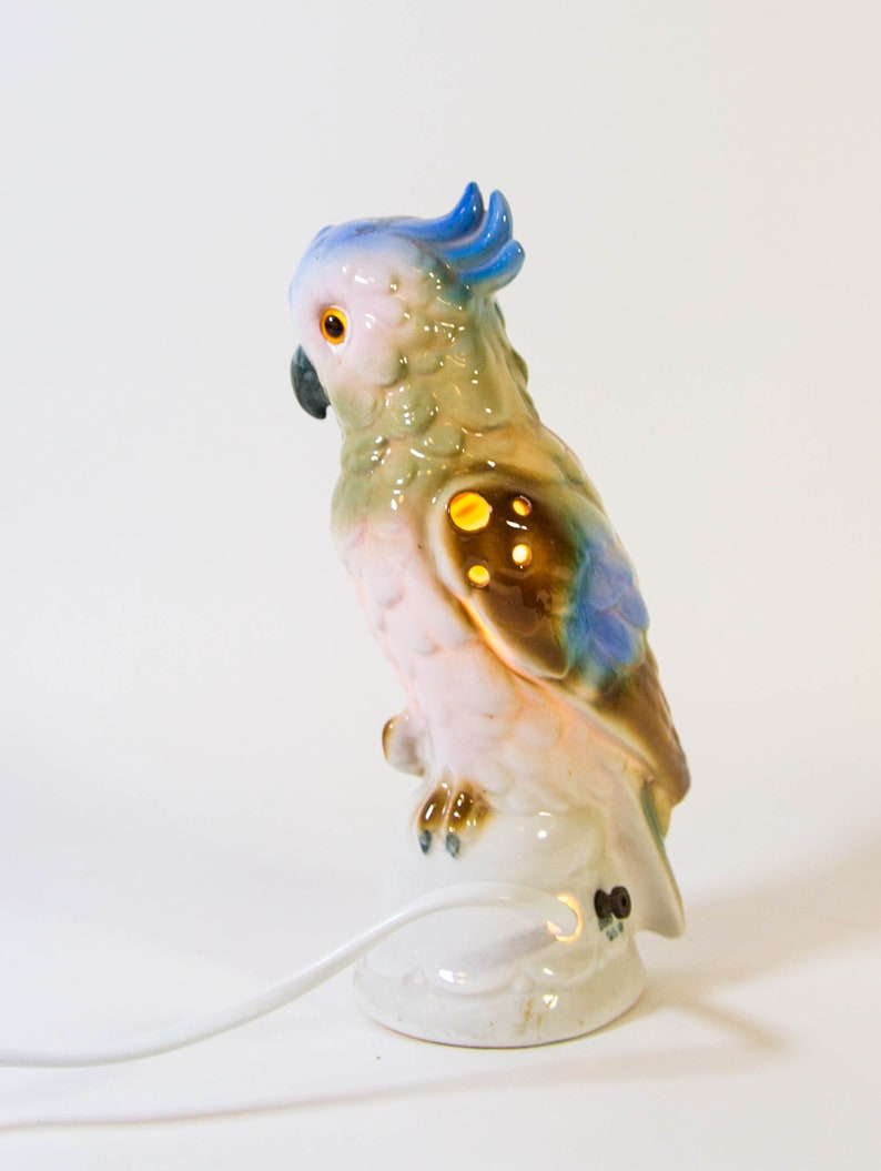 May include: A ceramic night light shaped like a parrot with blue, green, brown, and pink accents. The parrot is perched on a white base with a cord.