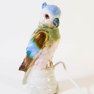 May include: A ceramic parrot figurine with blue, green, brown, and pink paint. The parrot is perched on a white base with a cord coming out of the base.