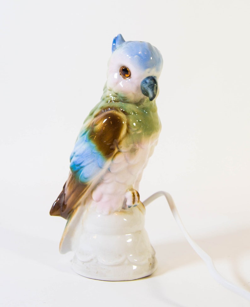 May include: A ceramic parrot figurine with blue, green, brown, and pink paint. The parrot is perched on a white base with a cord coming out of the base.