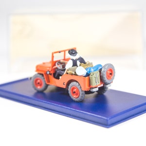 May include: A red toy jeep with a figure in the driver's seat. The jeep has a blue base and is on a blue display stand.