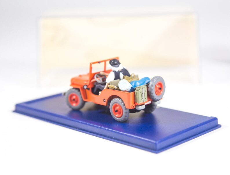 May include: A red toy jeep with a figure in the driver's seat. The jeep has a blue base and is on a blue display stand.
