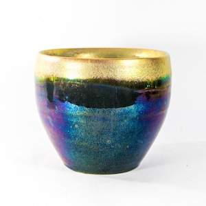 Könnte beinhalten: Ein Keramikgefäß mit goldenem Rand und einem Körper, der von Dunkelblau zu schillerndem Lila und Grün übergeht. Die handgefertigte Keramik hat eine einzigartige, strukturierte Oberfläche und eine weite Öffnung.