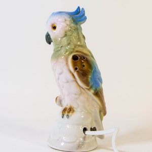 May include: A ceramic parrot nightlight with a blue crest, green and brown body, and pink underbelly. The parrot is perched on a white base with a cord.