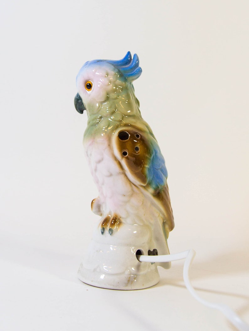 May include: A ceramic parrot nightlight with a blue crest, green and brown body, and pink underbelly. The parrot is perched on a white base with a cord.