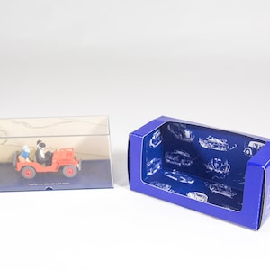 May include: A red toy jeep with two figures inside. The jeep is in a clear plastic display case. The display case is on a blue base with white lettering. The display case is next to a blue cardboard box with a clear plastic window. The cardboard box has a blue background with white images of cars.