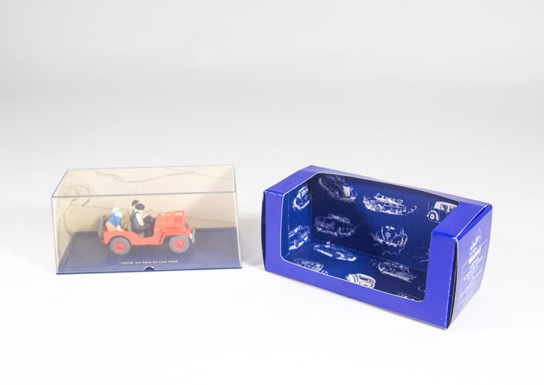May include: A red toy jeep with two figures inside. The jeep is in a clear plastic display case. The display case is on a blue base with white lettering. The display case is next to a blue cardboard box with a clear plastic window. The cardboard box has a blue background with white images of cars.