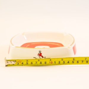 May include: White and red ceramic ashtray with gold trim. The ashtray has a rectangular shape with rounded corners. The text "Born 1820" and a figure in a red coat are visible. The ashtray measures approximately 4 inches in length.