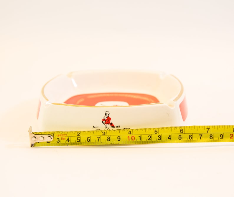 May include: White and red ceramic ashtray with gold trim. The ashtray has a rectangular shape with rounded corners. The text "Born 1820" and a figure in a red coat are visible. The ashtray measures approximately 4 inches in length.