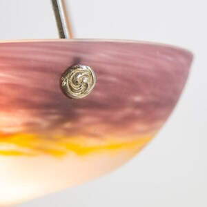 Pu&ograve; includere: Primo piano di un paralume decorativo in vetro con una sfumatura di colore, che passa dal viola scuro in alto al giallo vibrante in basso. Un bottone argentato ornato &egrave; fissato sul lato.