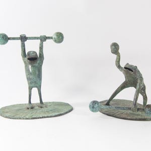 May include: Two green frog sculptures, one lifting a barbell and the other lifting a dumbbell, both standing on a leaf-shaped base.