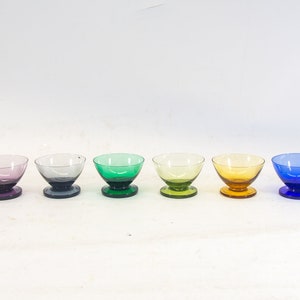 Op de afbeelding: Zes kleine glazen kommen met voetjes in verschillende kleuren, waaronder paars, groen, geel en blauw.