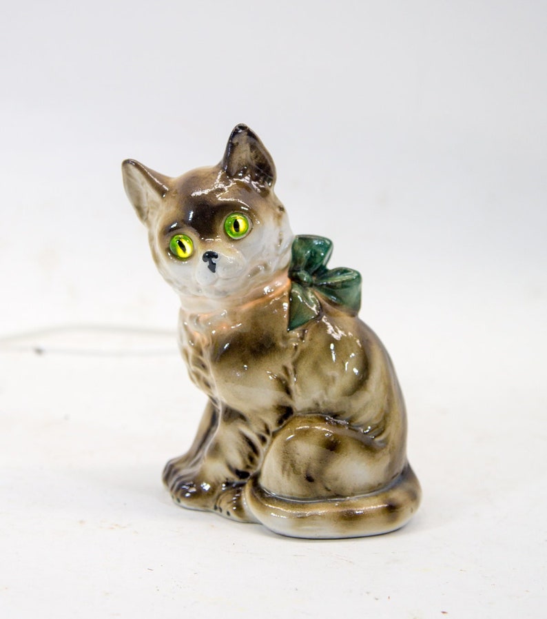 May include: A ceramic cat figurine with green eyes and a green bow. The cat is sitting with its tail curled around its legs. The figurine is painted in shades of brown and white.