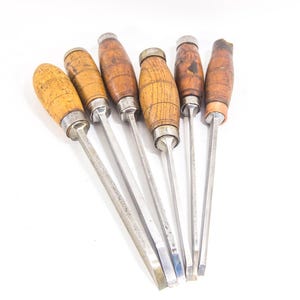 May include: Six wood carving chisels with wooden handles. The chisels are arranged in a fan shape with the handles pointing towards the top of the image. The blades are silver and the handles are a light brown colour.