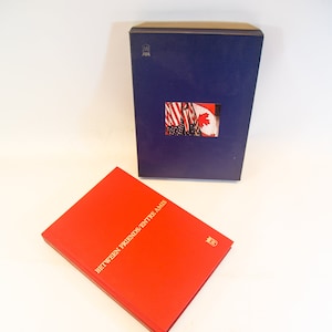 Puede incluir: Dos libros de tapa dura con cubiertas roja y azul. El libro rojo tiene el título "Between Friends/Entre Amis" en letras doradas. El libro azul tiene una pequeña imagen de las banderas estadounidense y canadiense.