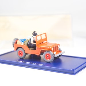 May include: A red toy jeep with two figures inside. The jeep is on a blue display base with the words 'Tintin Au Pays De L'or Noir' printed on it.
