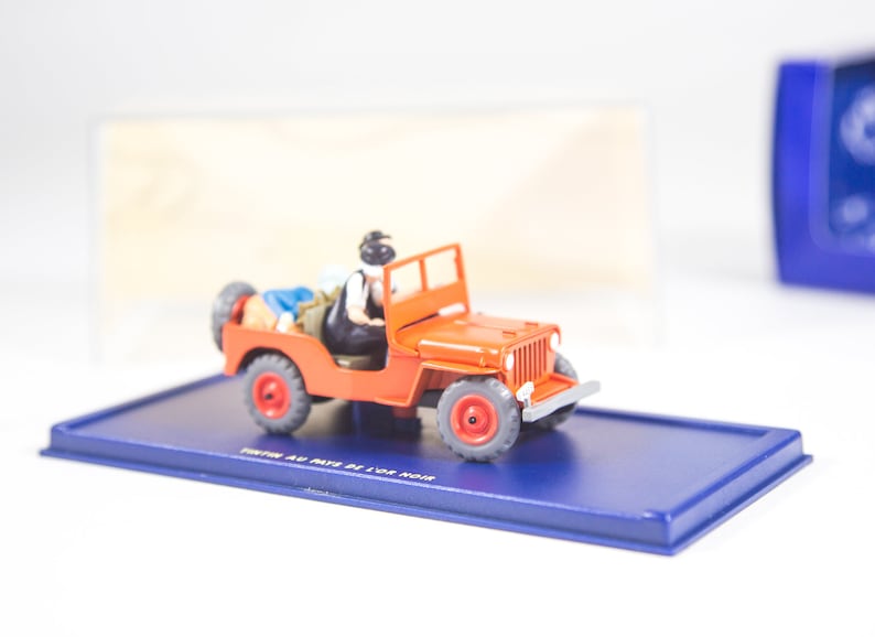 May include: A red toy jeep with two figures inside. The jeep is on a blue display base with the words 'Tintin Au Pays De L'or Noir' printed on it.