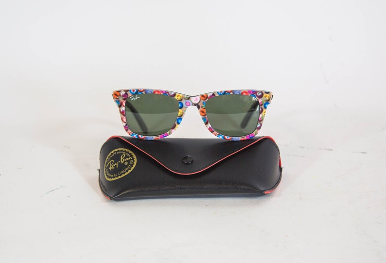 Puede incluir: Gafas de sol Ray-Ban con montura de patr&oacute;n de bolas de billar multicolor y lentes verde oscuro, acompa&ntilde;adas de un estuche protector negro con el logotipo de Ray-Ban dorado impreso en la solapa frontal y cierre de bot&oacute;n a presi&oacute;n.