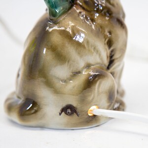 May include: A ceramic figurine of a brown and white dog with a green tail, sitting with its head turned to the side. The figurine has a light bulb in the back and a cord coming out of the bottom.