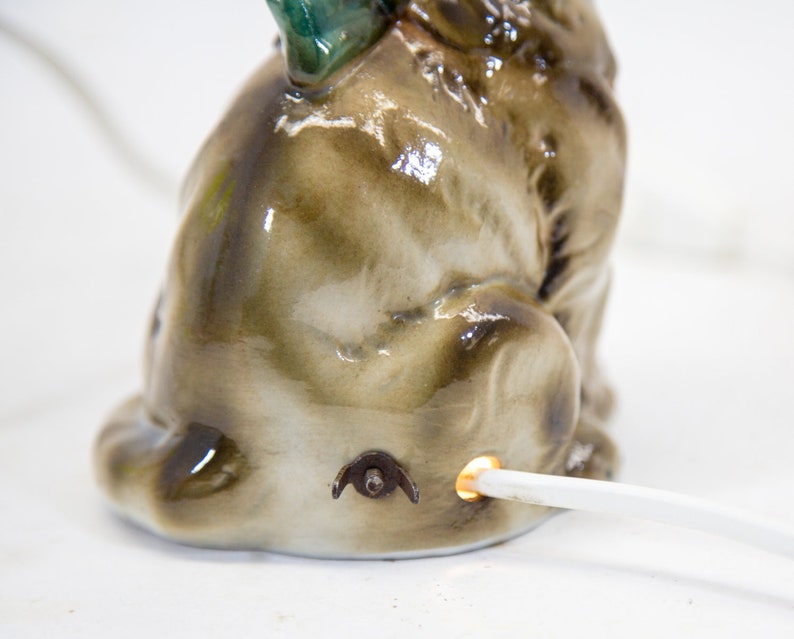 May include: A ceramic figurine of a brown and white dog with a green tail, sitting with its head turned to the side. The figurine has a light bulb in the back and a cord coming out of the bottom.