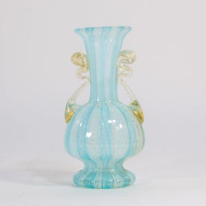 May include: A decorative glass vase with a light blue and white striped pattern. The vase has two gold-colored handles and a flared top. The vase is approximately 8 inches tall.