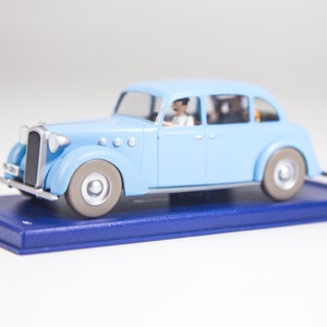 May include: A light blue toy car with a silver roof and a driver and passengers inside. The car is on a blue display base.