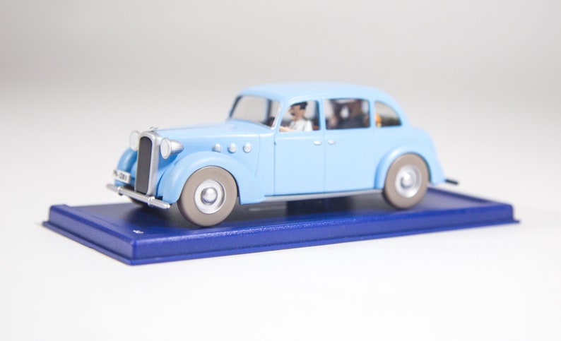 May include: A light blue toy car with a silver roof and a driver and passengers inside. The car is on a blue display base.