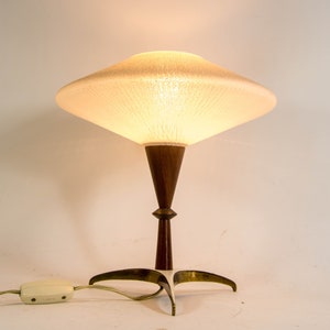 Könnte beinhalten: Eine Tischlampe im Retro-Stil mit einem konischen, hellbeigen Lampenschirm. Der Sockel besteht aus dunklem Holz und Messing. Die Lampe hat einen dreibeinigen Fuß und ein Kabel mit Schalter. Die Lampe ist eingeschaltet und strahlt ein warmes Licht aus.