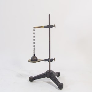 May include: An antique, black metal balance scale with a three-legged base. A small brass arm extends from the vertical support, holding a chain and a spherical weight. The scale is designed for scientific or decorative purposes.