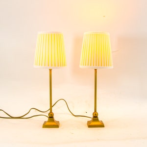 Op de afbeelding: Twee gouden tafellampen met witte lampenkappen met gele strepen. De lampen hebben een ronde voet en een vierkante voet.