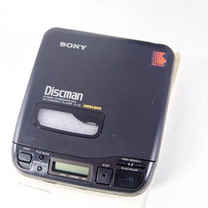 May include: A black Sony Discman CD player with the text "Discman 8 Times Oversampling CD Compact Player D-32 MEGA BASS" on the front. The player has a silver display screen and buttons for open, play mode, repeat/enter, stop, AMS/search, and play/pause.