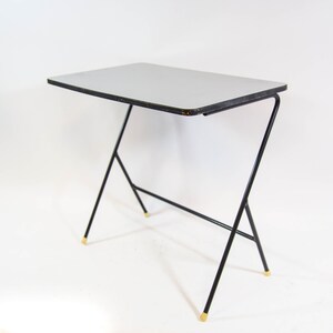 May include: A small, black metal table with a light grey top. The table has a simple, modern design with a Z-shaped base and gold-coloured accents.
