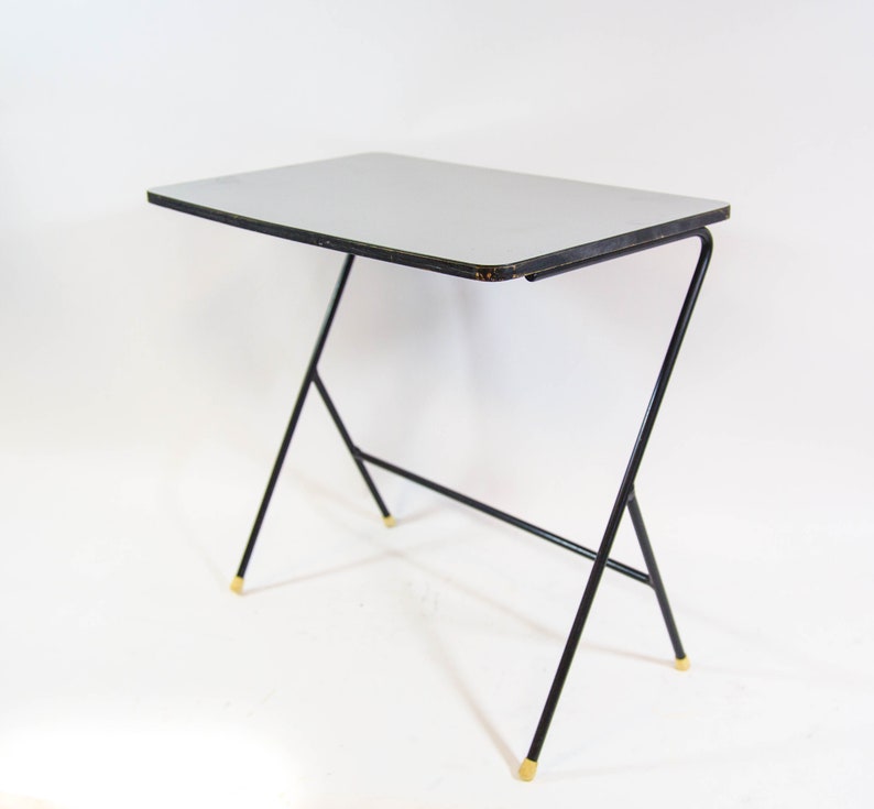 May include: A small, black metal table with a light grey top. The table has a simple, modern design with a Z-shaped base and gold-coloured accents.