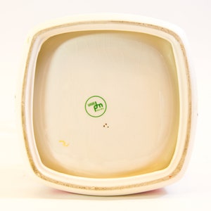 May include: Cream-colored ceramic container with a square shape and rounded corners. The interior is off-white with a green circular stamp that reads "WOODS ENGLAND". The rim is a light beige color.
