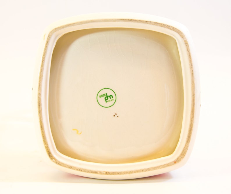 May include: Cream-colored ceramic container with a square shape and rounded corners. The interior is off-white with a green circular stamp that reads "WOODS ENGLAND". The rim is a light beige color.