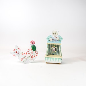 May include: Two whimsical wooden figurines. One is a white cat with red stripes and a mouse with a pink rose on its back. The other is a puppet show with a figure of a woman and two puppets on a stage.