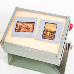 Puede incluir: Visor de diapositivas vintage con dos diapositivas iluminadas desde el interior. El visor es de metal gris y tiene una luz roja en el lateral. Las diapositivas muestran imágenes de personas. La marca "hamafix" es visible.