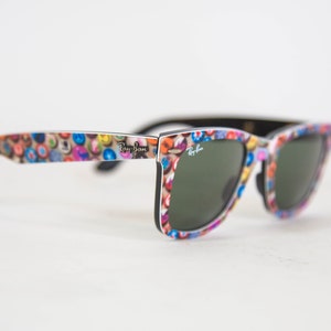 Puede incluir: Gafas de sol Ray-Ban con un patr&oacute;n multicolor de peque&ntilde;os iconos circulares en las monturas. Las lentes son de color verde oscuro y el logotipo de Ray-Ban est&aacute; impreso en blanco en la lente derecha y en negro en la patilla exterior.