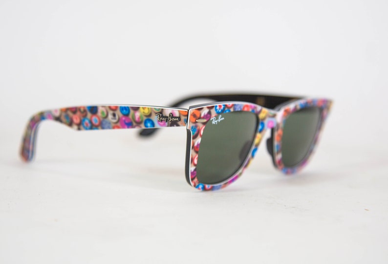 Puede incluir: Gafas de sol Ray-Ban con un patr&oacute;n multicolor de peque&ntilde;os iconos circulares en las monturas. Las lentes son de color verde oscuro y el logotipo de Ray-Ban est&aacute; impreso en blanco en la lente derecha y en negro en la patilla exterior.
