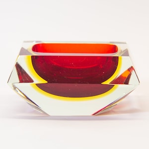 Op de afbeelding: Een asbak van rood en geel glas met een geometrisch ontwerp. De asbak is gemaakt van helder glas met een rood en geel interieur.