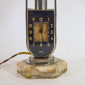 Peut inclure: Une lampe-horloge en argent et noir avec une base en marbre. Le cadran de l'horloge a des chiffres romains noirs et une aiguille des heures et des minutes dor&eacute;e. Le cadran de l'horloge porte &eacute;galement l'inscription "MOFEM".