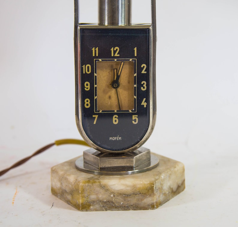 Peut inclure: Une lampe-horloge en argent et noir avec une base en marbre. Le cadran de l'horloge a des chiffres romains noirs et une aiguille des heures et des minutes dor&eacute;e. Le cadran de l'horloge porte &eacute;galement l'inscription "MOFEM".
