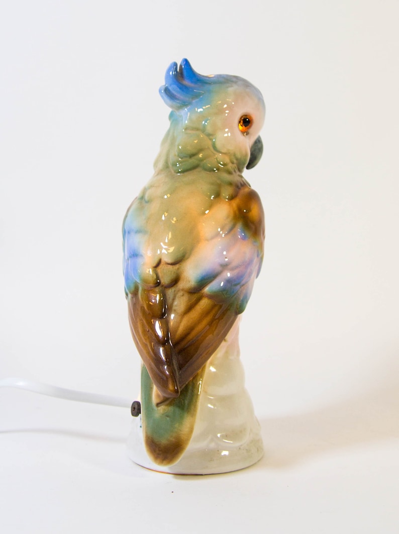 May include: A ceramic parrot lamp with a blue, green, and brown color scheme. The parrot is perched on a branch and has a white base.
