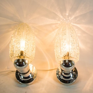 Puede incluir: Dos apliques de pared con pantallas de vidrio texturizado y bases de metal cromado. Los apliques están encendidos, proyectando una luz cálida y patrones intrincados en la pared. Una lámpara vintage.