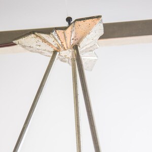 Pu&ograve; includere: Primo piano di un lampadario vintage in stile Art D&eacute;co. L'apparecchio presenta una parte superiore argentata a forma di stella con tre sottili aste argentate che si estendono verso il basso. Il design ricorda gli anni '20 e '30.